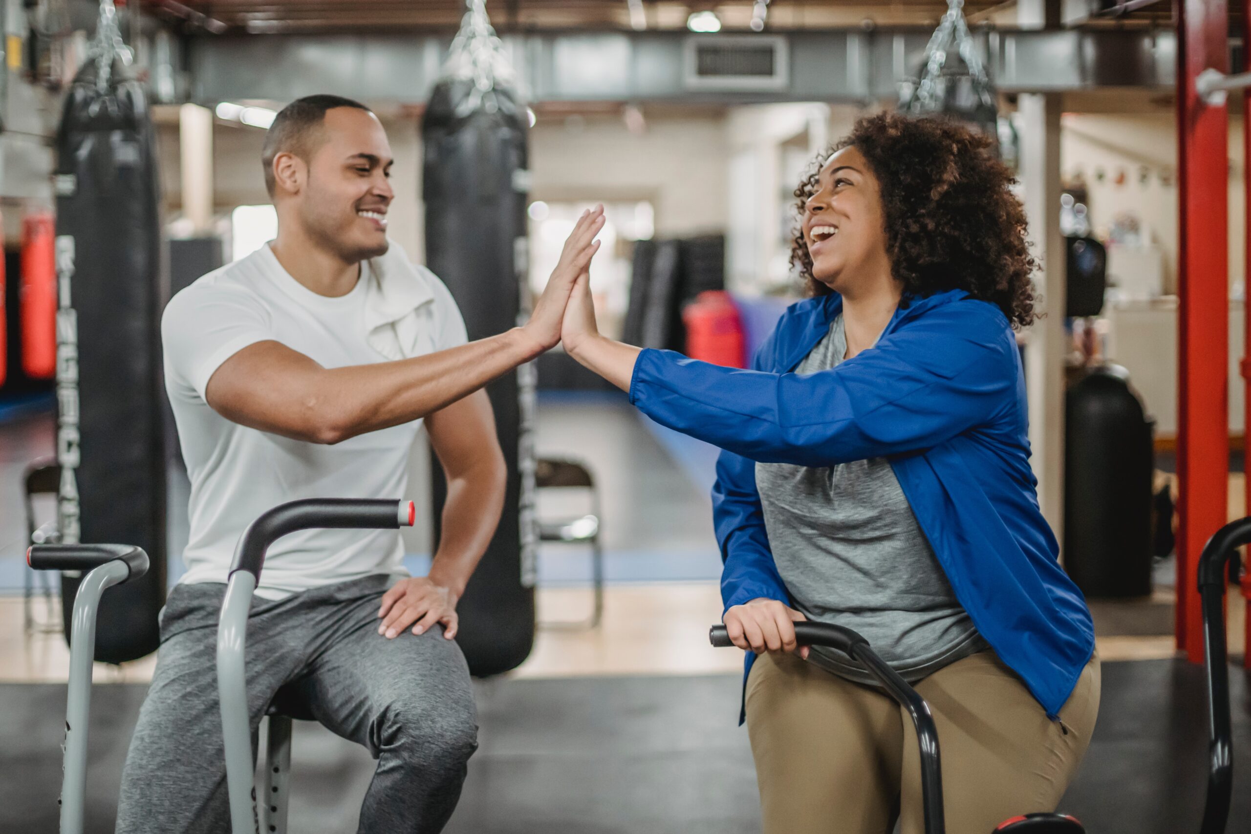 A man and a woman smiling and giving each other a high-five in a gym, with the woman sitting on an exercise bike and the man standing beside her.